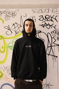 a man in a black hoodie standing in front of a wall covered in graffiti
